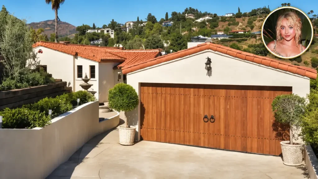 Sabrina Carpenter House featuring a Spanish Colonial Revival exterior, terracotta roof, wooden garage door, and hillside landscaping.