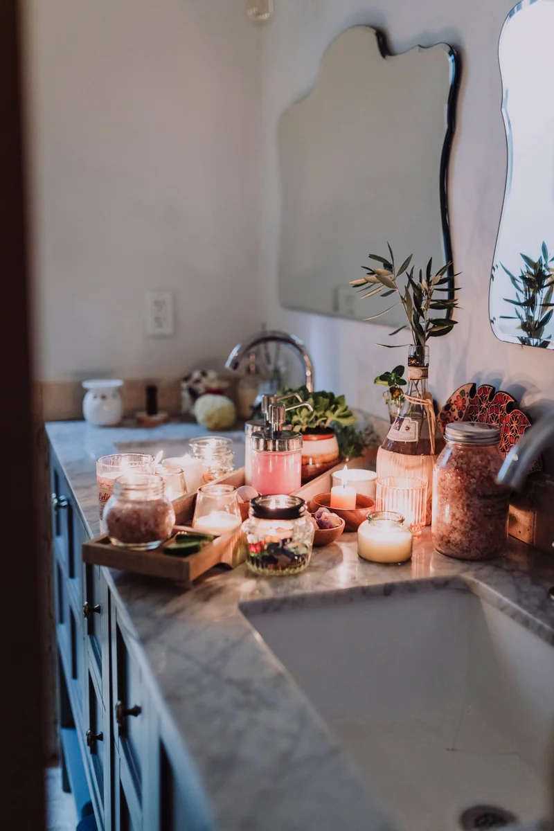 A Small Room, Dressed for December, That Still Works Every Morning 6 Lit candles in jars on a bathroom counter beside a tray, soap dispenser and greenery