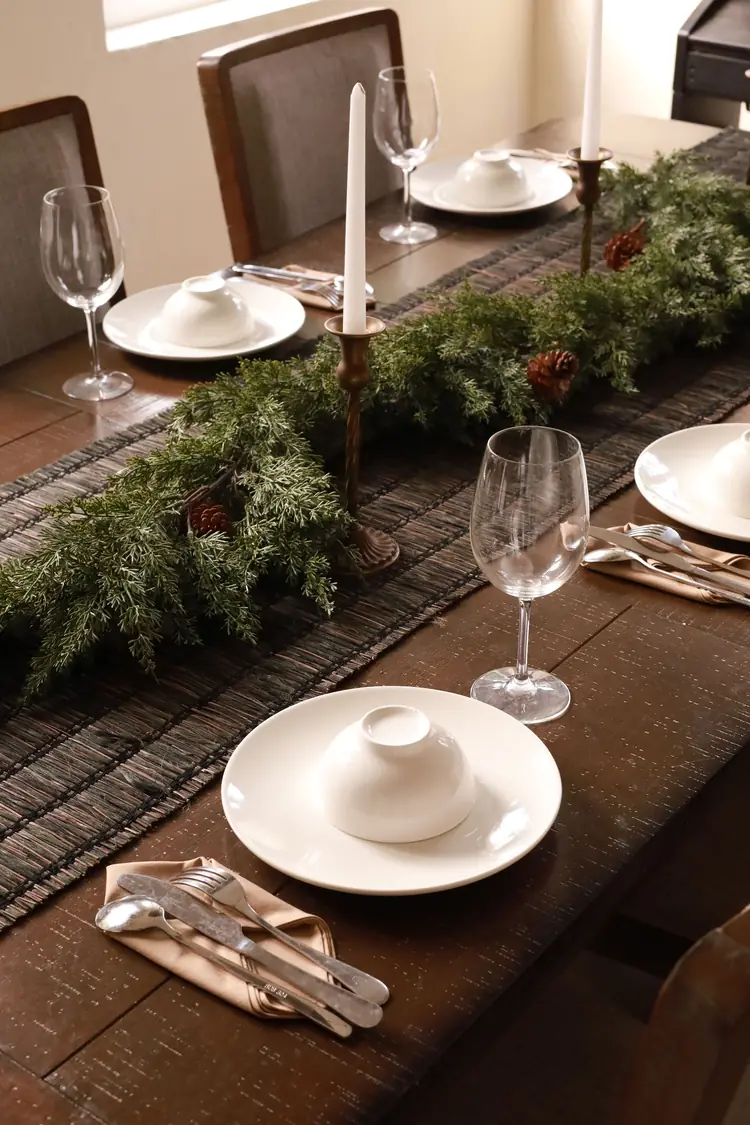 Seven Moves That Turn the Dining Table Into Christmas 1 A dark woven runner with a green garland and tall white candles down the middle of a set dining table