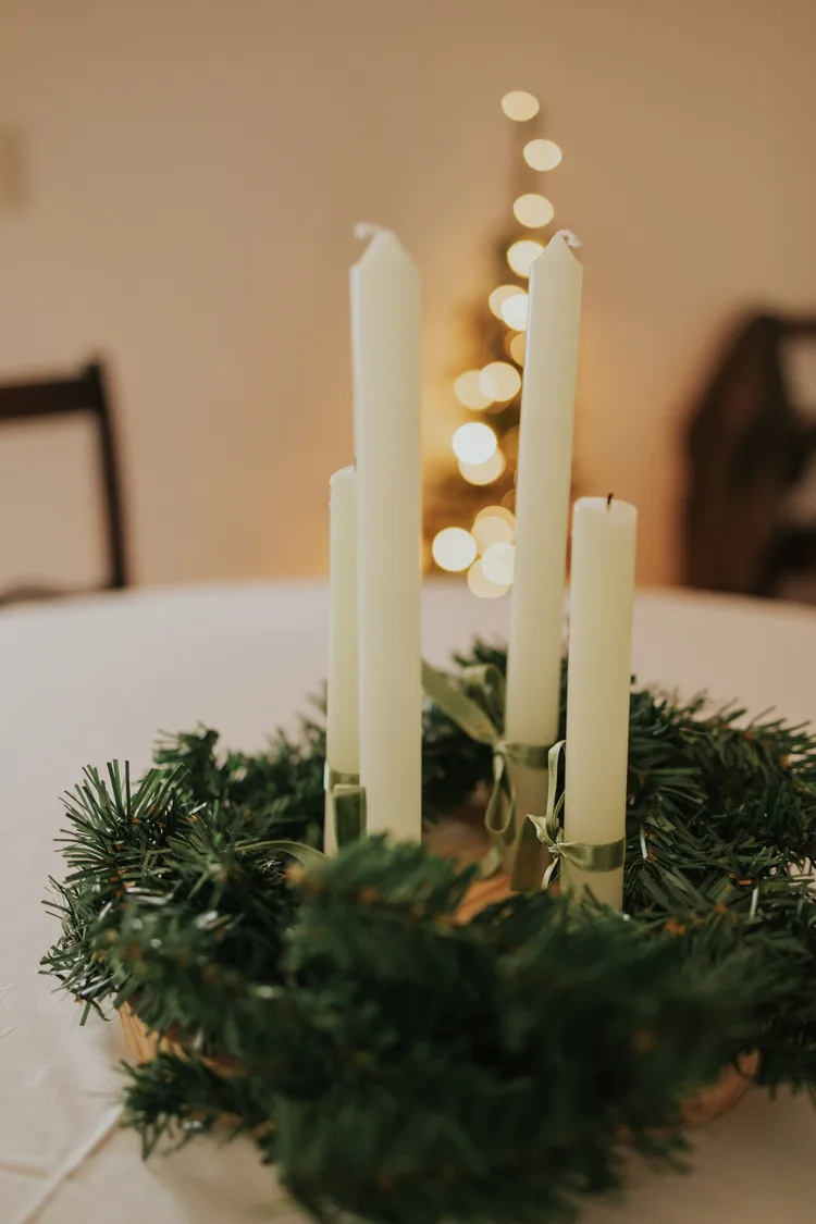 Seven Moves That Turn the Dining Table Into Christmas 2 Four tall white candles in a green evergreen wreath on a white tablecloth, with a lit Christmas tree behind