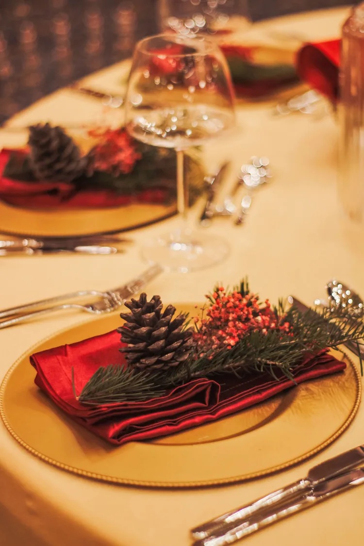Seven Moves That Turn the Dining Table Into Christmas 3 Christmas place settings with gold chargers, red napkins, pinecones and wine glasses on a cream tablecloth