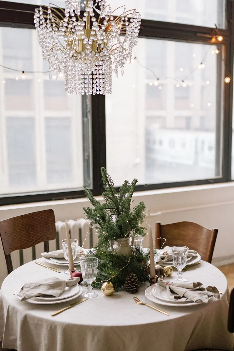 Seven Moves That Turn the Dining Table Into Christmas 7 A small round table with a cream cloth set for Christmas dinner under a crystal chandelier