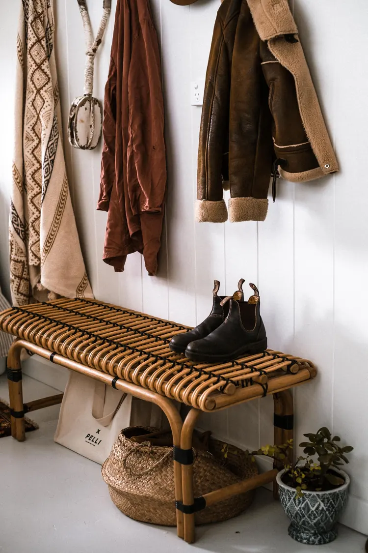 A rattan entryway bench with boots, coats on hooks above, and a basket underneath