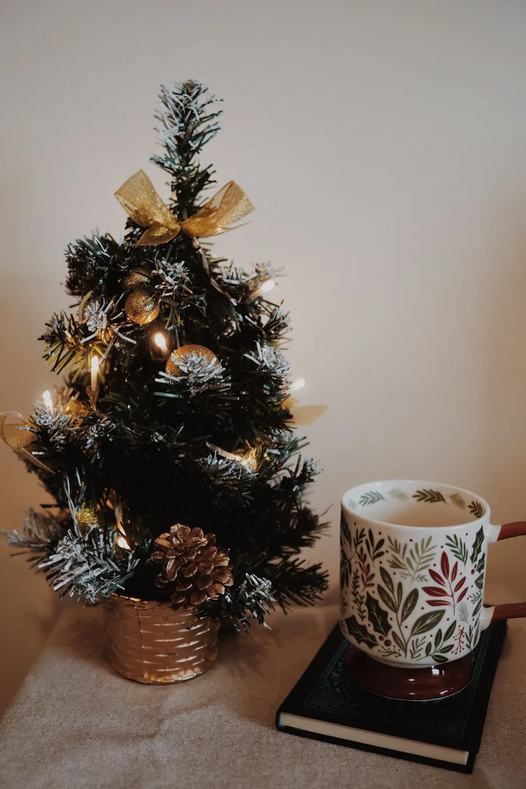 A small frosted Christmas tree with warm lights in a gold pot beside a festive mug