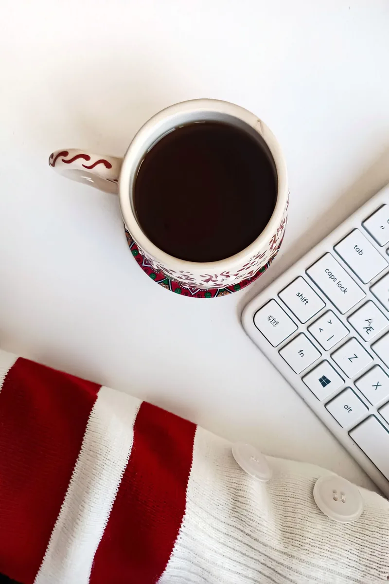 A festive patterned mug of coffee on a mat beside a keyboard on a clear white desk