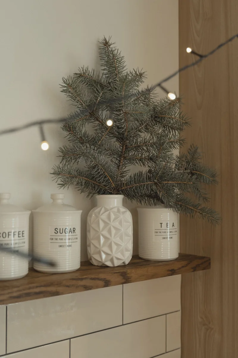 Fresh fir sprigs in a white ceramic jar on a wooden shelf with string lights