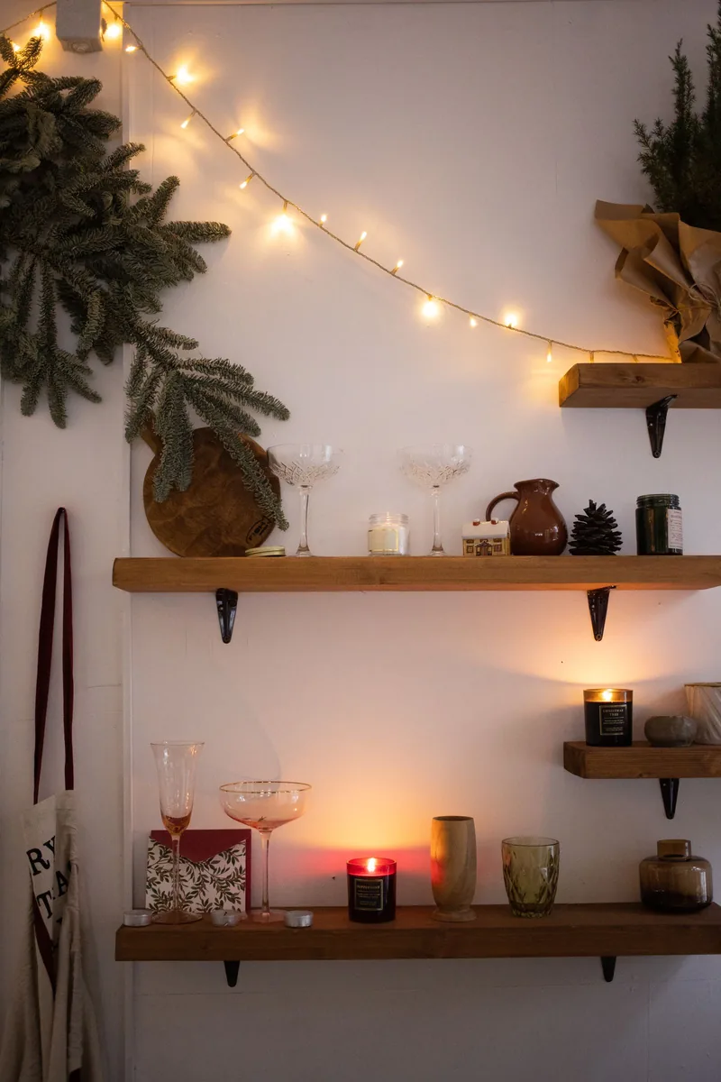 Warm string lights draped along wooden wall shelves with candles and pine branches