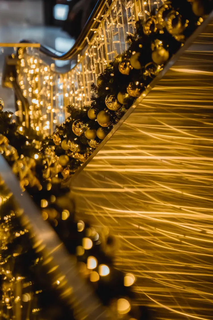 A green garland with gold baubles and warm lights along a metal railing