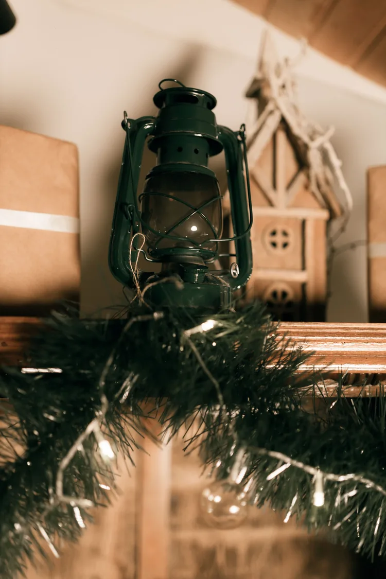 A dark green vintage lantern on a shelf behind a Christmas garland with warm lights