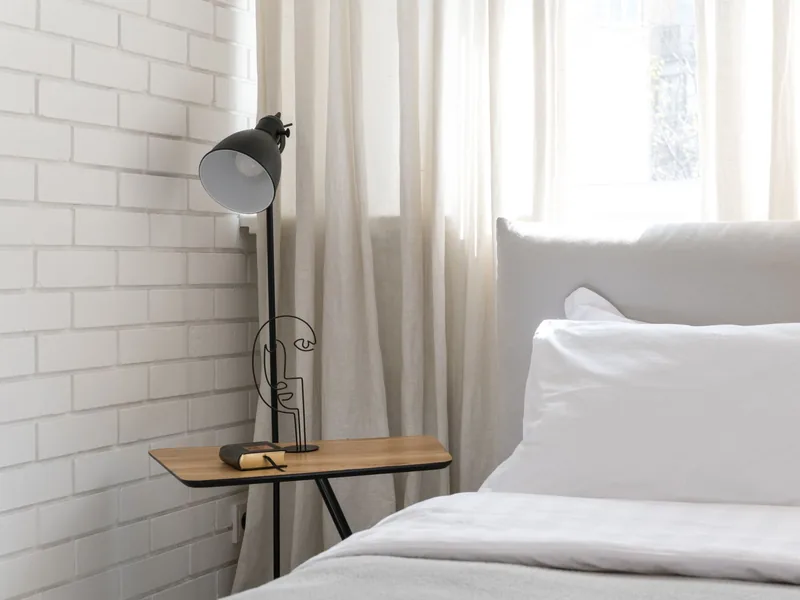 A Calm, Green Holiday Bedroom You Can Actually Sleep In 7 A tidy bedroom with white bedding, one small bedside table and a floor lamp beside a white brick wall