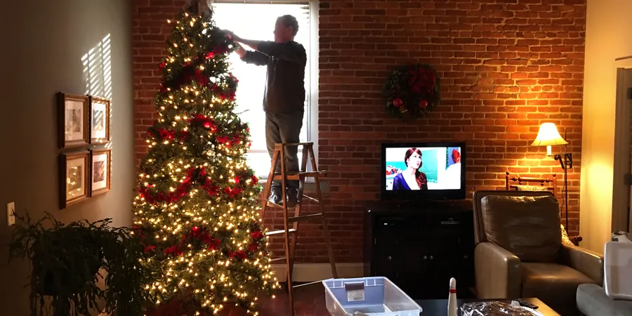 A Warm, Lived In Holiday Room You Can Finish This Weekend 2 A tall Christmas tree being decorated beside a living room window, showing a corner tree placement that keeps the floor clear.