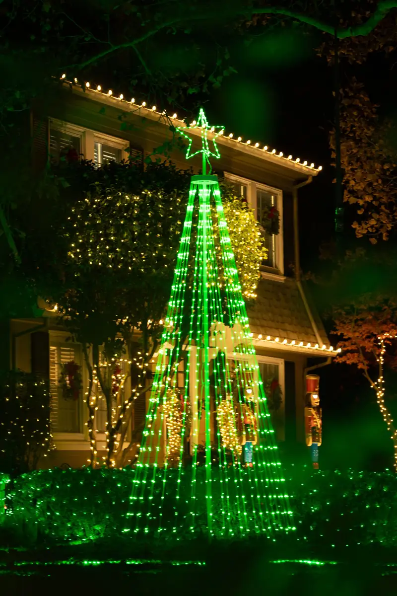 A house front at night with warm string lights along the roofline, a tall green light display with a star in the yard, and wreaths in the windows.