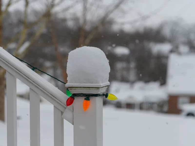 A snow-covered porch railing post with a short strand of multicolour Christmas lights wrapped around it.