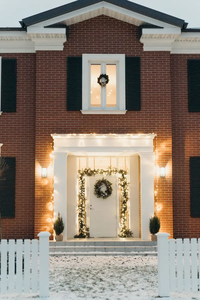 A brick house entrance at dusk with a wreath on the door, garland framing the doorway, and warm string lights along the porch.