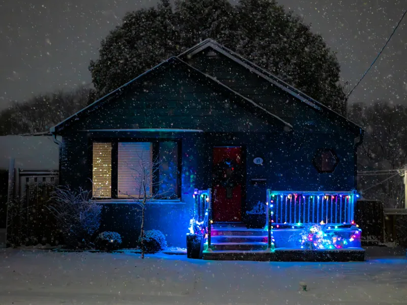A house in falling snow at night with multicolour lights along the porch railing and glowing figures in the yard.